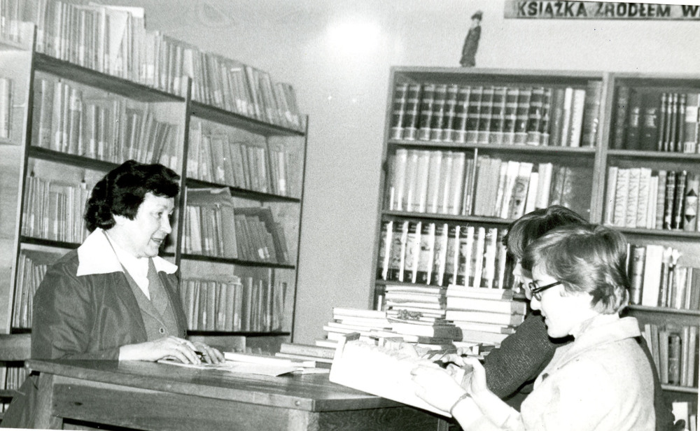 Bibliotekarka siedzi przy soliku w czytelni i udziela informacji dwóm czytelniczkom.
