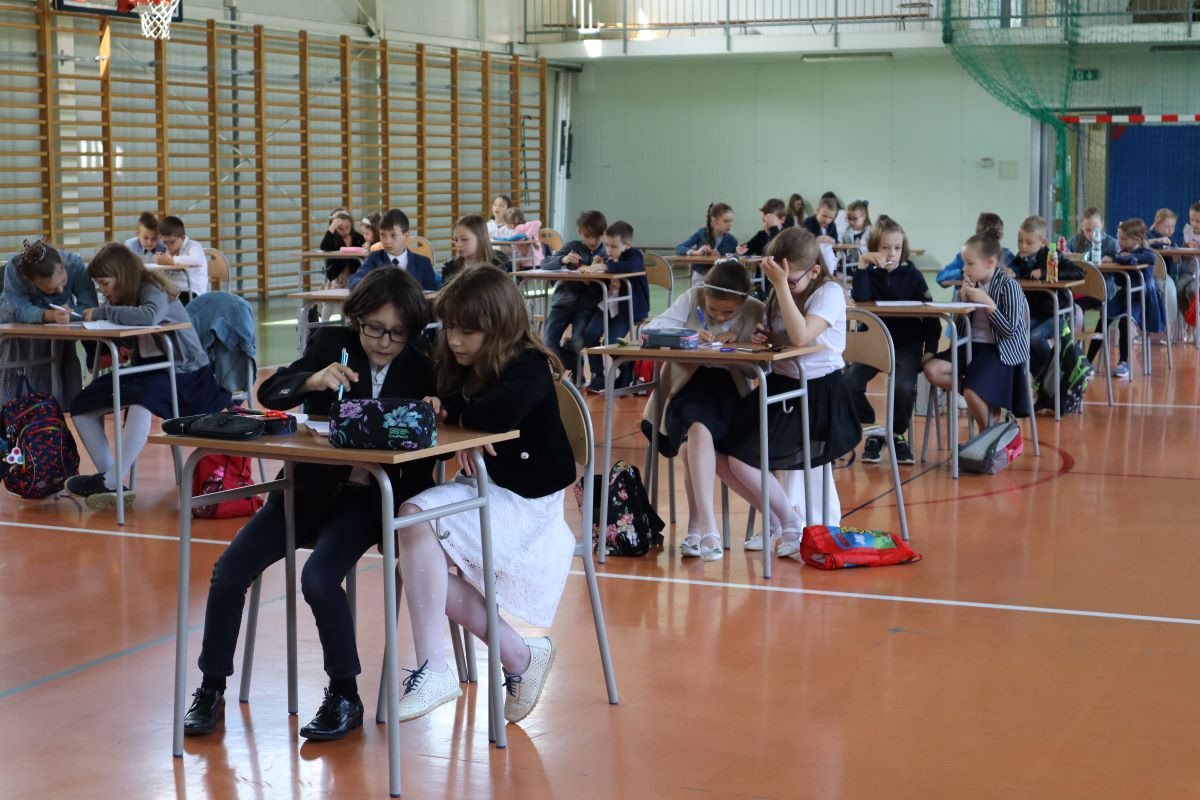 Uczniowie siądzą przy stolikach i rozwiązują test finałowy.